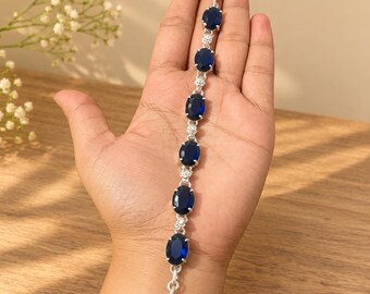 May include: A silver bracelet featuring six oval-shaped deep blue gemstones arranged in a line, separated by small clear stones. The bracelet includes a toggle clasp closure at one end and a circular ring at the other for secure fastening.