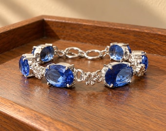 May include: A silver-toned bracelet featuring multiple oval-shaped, deep blue faceted stones. The stones are linked together by small silver-toned metal flower-shaped connectors and circular chain links, creating a continuous, articulated design.
