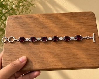 May include: A silver bracelet featuring six oval-cut deep red garnet stones alternating with small floral-shaped clusters of clear white stones. The piece is finished with a toggle clasp closure, consisting of a circular ring and a T-bar component.