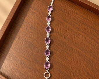 May include: A silver bracelet featuring six oval-cut light purple gemstones arranged in a vertical line, connected by small floral-shaped silver links, and finished with a toggle clasp consisting of a T-bar and a circular ring at the end.