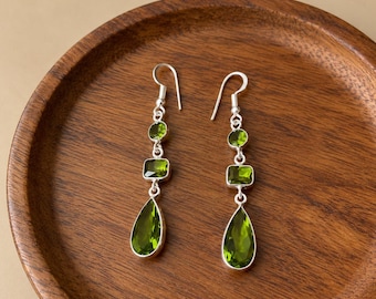 May include: A pair of silver dangle earrings featuring three green faceted stones in each piece. The stones are arranged in a vertical line, consisting of a small round stone, a rectangular stone, and a large teardrop-shaped stone at the base.