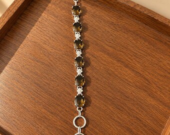 May include: A silver bracelet featuring six oval-cut smoky quartz gemstones alternating with small floral-shaped silver links. The bracelet includes a toggle clasp closure with two circular rings at one end and a T-bar at the other end.