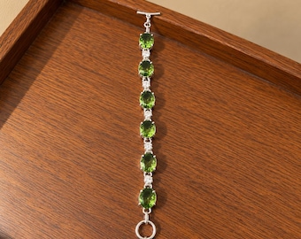 May include: A silver-toned bracelet featuring six oval-cut vibrant green stones alternating with small clear round stones. The piece is finished with a toggle clasp closure at the top and two circular rings at the bottom end.