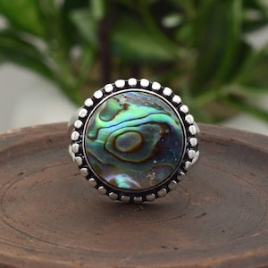 Könnte beinhalten: Ein Silberring mit einem großen, runden Abalone-Muschel-Mittelteil. Die Muschel zeigt schillernde Blau-, Grün- und Lilatöne. Der Ring hat einen mit Perlen besetzten Rand. Der Ring liegt auf einer Holzoberfläche.