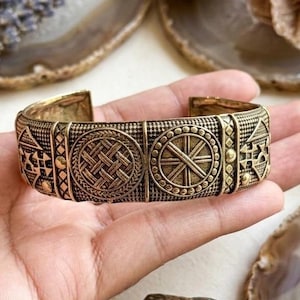 May include: A wide brass cuff bracelet featuring a series of repeating geometric patterns, including circular motifs with woven and radial line designs, separated by vertical textured panels with small raised dots in a metallic gold finish.