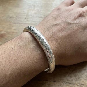 Könnte beinhalten: Ein Armreif aus silberfarbenem Metall mit einem detaillierten, geprägten Blumen- und Blattmuster, das die gesamte Außenfläche bedeckt. Der Armreif hat ein abgerundetes, poliertes Profil und ein durchgehendes Design.