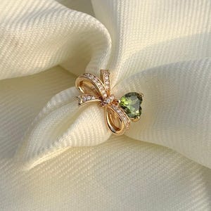 Könnte beinhalten: Ein goldfarbener Ring mit einem Schleifendesign, besetzt mit kleinen Diamanten und einem herzförmigen grünen Edelstein. Der Ring ist vor einem weißen Stoffhintergrund platziert, der die Details des Schmucks hervorhebt.