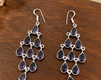 May include: A pair of silver chandelier earrings featuring multiple teardrop-shaped purple amethyst stones arranged in a tiered, cascading design. Each earring hangs from a simple silver hook and displays a series of faceted dark purple gems.