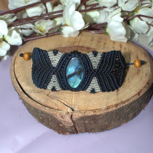 Könnte beinhalten: Handgefertigtes Makramee-Armband mit einem ovalen Labradorit-Stein in Blau- und Grüntönen. Das Armband ist schwarz und beige, mit Holzperlen an jedem Ende. Präsentiert auf einer Holzscheibe.