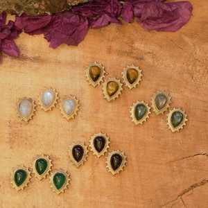 Könnte beinhalten: Eine Sammlung von Ohrsteckern mit Edelsteinen in verschiedenen Farben, darunter Grün, Lila, Gelb und Weiß. Jeder Ohrring hat einen tropfenförmigen Stein, der in einen goldfarbenen, sonnenförmigen Rahmen gefasst ist. Die Ohrringe liegen auf einer Holzoberfläche.
