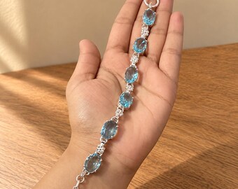 May include: A silver bracelet featuring six oval-cut light blue gemstones arranged in a row, separated by small clusters of clear white stones. The piece is finished with a circular toggle clasp at one end and a T-bar closure at the other.