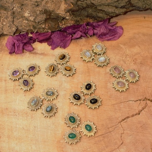 Könnte beinhalten: Eine Sammlung von Ohrringen mit Edelsteinen und goldfarbenen Fassungen. Die Ohrringe zeigen verschiedene Edelsteine, darunter Amethyst, Tigerauge, Mondstein, Rosenquarz und schwarzen Onyx. Sie sind auf einer Holzoberfläche mit lila Blütenblättern angeordnet.