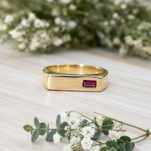 May include: A gold band ring featuring a single rectangular baguette-cut deep pink gemstone set horizontally into the flat top surface. The ring has a polished metallic finish and a smooth, wide band design that tapers slightly toward the back.