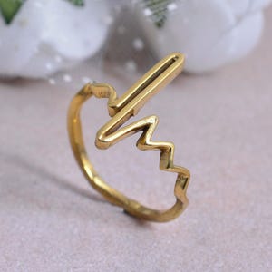 Könnte beinhalten: Ein goldfarbener Ring mit einem einzigartigen Design. Der Ring zeigt ein stilisiertes Herzschlag- oder Schallwellenmuster mit einem geraden Balken und einer Zickzacklinie. Der Ring besteht aus einem glänzenden, metallischen Material.