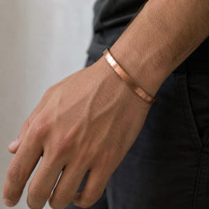 Könnte beinhalten: Ein glattes, poliertes Kupfer-Armband in einer einfachen, offenen Form. Das Metall hat einen warmen, rotbraunen metallischen Glanz und eine flache, gleichmäßige Oberfläche, die sich um das Handgelenk legt und eine klare Optik aufweist.