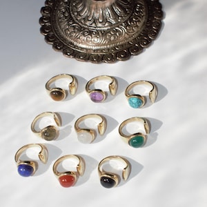 Könnte beinhalten: Eine Sammlung goldfarbener Ringe, jeder mit einem andersfarbigen Edelstein. Zu den Edelsteinen gehören Braun, Lila, Türkis, Grün, Blau, Rot und Schwarz. Die Ringe werden auf einer weißen Oberfläche präsentiert.