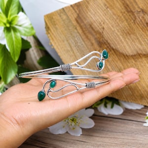 May include: A silver cuff bracelet featuring an open-ended design with swirling metal wire details and four teardrop-shaped green stones. The bracelet includes textured, wrapped metal accents near the ends where the green stones are set.
