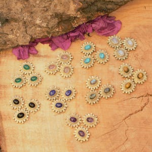 Könnte beinhalten: Eine Auswahl goldfarbener, sonnenförmiger Anhänger mit verschiedenen Edelsteinzentren, darunter Türkis, Amethyst und Labradorit. Die Anhänger sind auf einer Holzoberfläche mit einem Stück Rinde und lila Seidenpapier angeordnet.