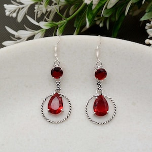 Könnte beinhalten: Silberne Ohrhänger mit roten Edelsteinen. Jeder Ohrring hat einen kleinen runden Edelstein, ein dekoratives Blumenelement, einen tropfenförmigen Edelstein und einen verdrehten Silberreif. Die Ohrringe hängen an einem Silberhaken.