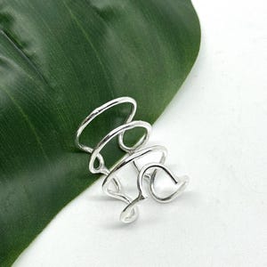 Könnte beinhalten: Ein silberfarbener Ring mit einem einzigartigen, offenen Design. Der Ring hat mehrere Bänder, die sich krümmen und schleifen und eine abstrakte, skulpturale Form bilden. Der Ring wird auf einem grünen Blatt vor weißem Hintergrund gezeigt.