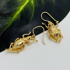 Könnte beinhalten: Ein Paar goldfarbene Käferohrringe. Die Ohrringe zeigen detaillierte Insektenmotive mit strukturierten Oberflächen und gebogenen Ohrhaken. Die Käfer sind mit ausgestreckten Beinen dargestellt.