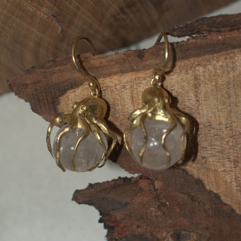 May include: A pair of gold-colored octopus earrings. Each earring features a small, round, light-colored stone held by the octopus's tentacles. The earrings have a hook closure.