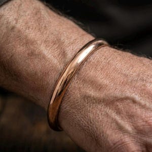 Könnte beinhalten: Ein poliertes Armband aus massivem Kupfer mit einer glatten, abgerundeten Oberfläche und einem durchgehenden Banddesign. Das Metall hat ein warmes, reflektierendes, rötlich-braunes Finish, das das Handgelenk umschließt und schlicht wirkt.