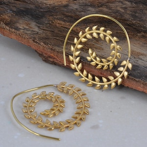 Könnte beinhalten: Zwei goldfarbene Spiralohrringe mit Blattdesign. Die Ohrringe sind aus Metall gefertigt und weisen ein filigranes, aufwendiges Muster auf. Die Blätter sind spiralförmig angeordnet und erzeugen einen einzigartigen und eleganten Look. Die Ohrringe werden auf einer Holzoberfläche präsentiert.