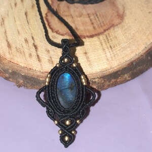 Könnte beinhalten: Eine handgefertigte Makramee-Halskette mit einem blauen Labradorit-Edelstein-Anhänger. Der Anhänger ist in schwarzem Makramee mit goldfarbenen Perlen gefasst. Die Halskette wird auf einer Holzoberfläche vor einem lila Hintergrund gezeigt.