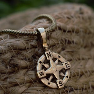 May include: A gold-coloured compass pendant with a star design at the centre, featuring the cardinal directions N, E, W, and S. The pendant hangs from a matching gold-coloured chain, set against a textured, woven background.