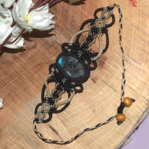 Könnte beinhalten: Handgefertigtes Makramee-Armband mit einem ovalen Labradorit-Stein in der Mitte. Das Armband ist aus schwarzen und beigefarbenen Kordeln gefertigt und mit kleinen goldfarbenen Perlen verziert. Es hat eine geflochtene Kordel mit Holzperlen am Ende.