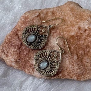 May include: A pair of ornate gold-colored earrings with a filigree design. Each earring features a central oval-shaped, light blue gemstone. The earrings have a hook closure and are displayed on a textured, tan-colored surface.