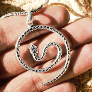 Peut inclure: Un collier pendentif argenté représentant un serpent enroulé. La tête du serpent est détaillée et le corps forme une spirale. Le pendentif est suspendu à une chaîne argentée. Le collier est présenté sur une main.