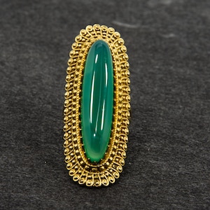 Könnte beinhalten: Ein goldfarbener Ring mit einem ovalen, hellgrünen Stein. Der Stein ist in einem detaillierten, verzierten Goldrahmen mit einem Perlenrand gefasst. Der Ring wird auf einer dunkelgrauen Oberfläche präsentiert.
