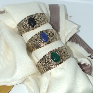 Può includere: Tre portatovaglioli argentati ornati, ciascuno con una pietra preziosa ovale di colore diverso: nero, blu e verde. Gli anelli hanno un motivo dettagliato e a spirale e sono esposti su un tovagliolo bianco piegato con strisce marroni.