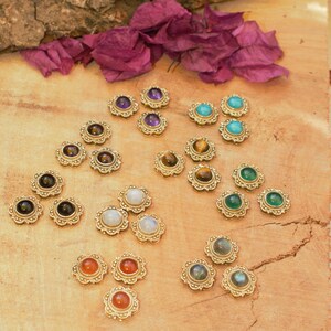 Könnte beinhalten: Eine Sammlung von Edelstein-Cabochons in goldfarbenen Fassungen. Die Edelsteine sind in verschiedenen Farben, darunter Lila, Türkis, Grün und Orange. Die Fassungen haben ein florales Design. Die Cabochons sind auf einer Holzoberfläche mit lila Blüten angeordnet.