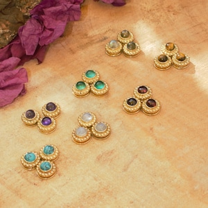 Könnte beinhalten: Eine Sammlung goldfarbener Schmuckstücke, jedes mit drei Edelsteinen. Die Edelsteine variieren in der Farbe, darunter Grün, Lila, Blau und Braun. Der Schmuck ist auf einer hellbraunen Holzoberfläche mit lila floralen Akzenten angeordnet.
