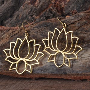 May include: A pair of gold-colored lotus flower earrings. The earrings are crafted in a cutout design, showcasing the intricate details of the flower's petals. The earrings have a hook closure and are displayed against a wooden background.