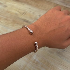 Könnte beinhalten: Ein offener Armreif in Roségold-Optik mit einem polierten, glatten Band, das an beiden Enden in einer kugelförmigen Verzierung endet. Jede Kugel ist mit drei horizontalen Gravurlinien versehen, was ein schlichtes und klares Design ergibt.