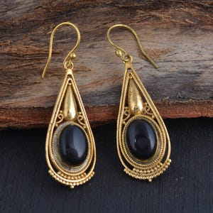May include: A pair of gold-toned drop earrings featuring an oval black stone set within a teardrop-shaped frame. The metal frame displays intricate wirework detailing and a smooth, polished finish, complemented by a curved hook earring wire.