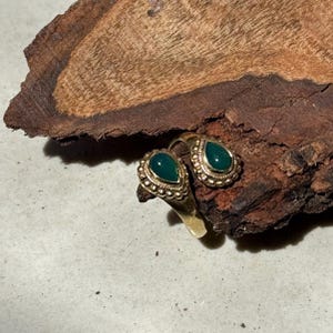 Könnte beinhalten: Ein goldfarbener Ring mit zwei tropfenförmigen grünen Edelsteinen. Der Ring hat ein detailliertes, strukturiertes Design um die Edelsteine. Der Ring wird auf einem Stück dunkelbraunem Holz präsentiert.