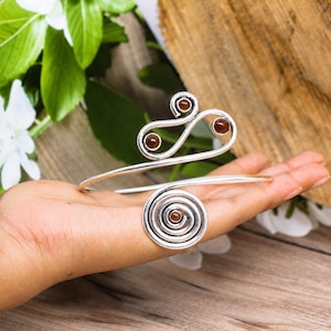 May include: A silver-toned metal arm cuff featuring a spiral design on one end and a double-loop shape on the other. The jewelry piece is accented with four small, round, dark amber-colored stones embedded within the metal swirls and loops.