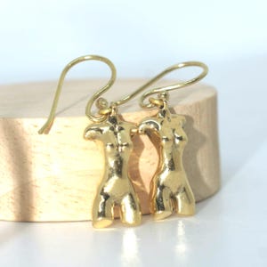Könnte beinhalten: Ein Paar goldfarbene Ohrringe mit einem stilisierten menschlichen Torso-Design. Die Ohrringe haben einen Hakenverschluss und werden auf einer hellen Holzoberfläche präsentiert. Die Ohrringe sind aus glänzendem Metall gefertigt.