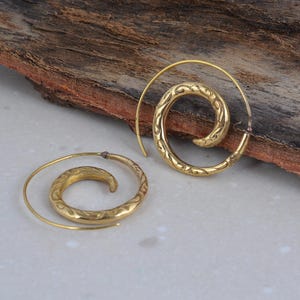 May include: A pair of spiral earrings in a gold tone. The earrings have an etched design on the inner spiral and a smooth, thin wire extending from the center. The earrings are displayed on a wooden surface.