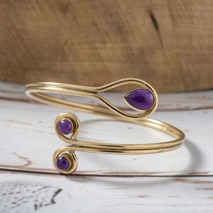Puede incluir: Un brazalete abierto de tono dorado con una piedra morada en forma de lágrima en un extremo y dos piedras moradas circulares más pequeñas en el otro. La banda metálica consta de dos alambres paralelos que se curvan formando bucles.