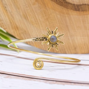 Puede incluir: Una pulsera de metal en tono dorado con un dije en forma de sol y una piedra redonda gris en el centro. La pulsera tiene un diseño abierto con un extremo en espiral y un motivo de rayos de sol texturizado con detalles intrincados.