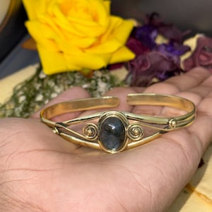 Könnte beinhalten: Ein goldfarbenes Metall-Armband mit einem zentralen, ovalen, dunkelgrauen Labradorit-Edelstein. Das Armband ist mit offenem Drahtgeflecht gestaltet und verfügt über zwei kleine dekorative Spiralen, die den Stein seitlich einrahmen.