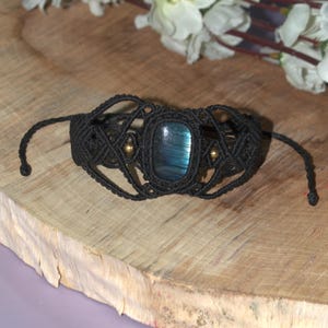 Könnte beinhalten: Ein schwarzes Makramee-Armband mit einem rechteckigen, irisierenden blauen Edelstein. Das Armband hat ein gewebtes Design mit kleinen goldfarbenen Perlen. Das Armband wird auf einer hellbraunen Holzoberfläche präsentiert.
