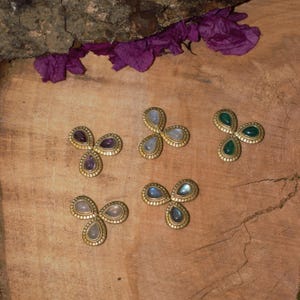 Könnte beinhalten: Fünf goldfarbene, dreiblättrige, blütenförmige Schmuckstücke mit verschiedenfarbigen Edelsteinen. Zu den Edelsteinen gehören Amethyst, Mondstein und Smaragd. Der Schmuck wird auf einer Holzoberfläche mit violetten Blütenblättern präsentiert.