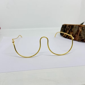 Könnte beinhalten: Eine goldfarbene, randlose Brille mit einem einzigartigen geschwungenen Steg. Jeder Gläserbereich hat einen kleinen, klaren Edelstein. Die Brille befindet sich vor einem weißen Hintergrund mit einem Holzstück im Hintergrund.